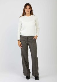 Giovane donna in piedi rivolta in avanti, indossa un maglione bianco, pantaloni grigi a gamba larga con risvolti, scarpe nere, con una mano in tasca.