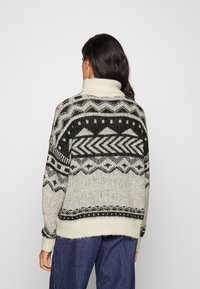 Stickad tröja med hög krage, ljusgrå bas med svarta geometriska mönster, samt ribbade ärmslut och nederkant. Texturerat, varmt tyg.