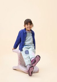 Kind in een blauwe jas en gestreepte tuinbroek met kleurrijke patches, zittend op een wit en roze gespikkeld krukje, met paarse schoenen.