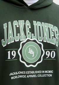 Sudadera verde con el texto "JACK & JONES" en blanco y verde claro, con un logo circular. Incluye el año "1990" y detalles de la marca.
