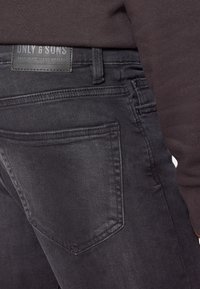Pantalones vaqueros de denim negro con un acabado texturizado, que presentan un bolsillo trasero rectangular y una etiqueta con la inscripción "ONLY & SONS" cosida en la cinturilla.