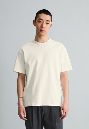 Jeune homme aux cheveux courts portant un t-shirt blanc uni, un pantalon noir, une chaîne en argent et un bracelet, debout devant un fond clair uni.