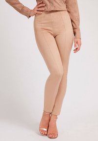 Leggings nude in suede sintetico con vita alta, texture liscia e dettagli di cucitura lungo la parte anteriore; abbinati a tacchi nude con cinturini.