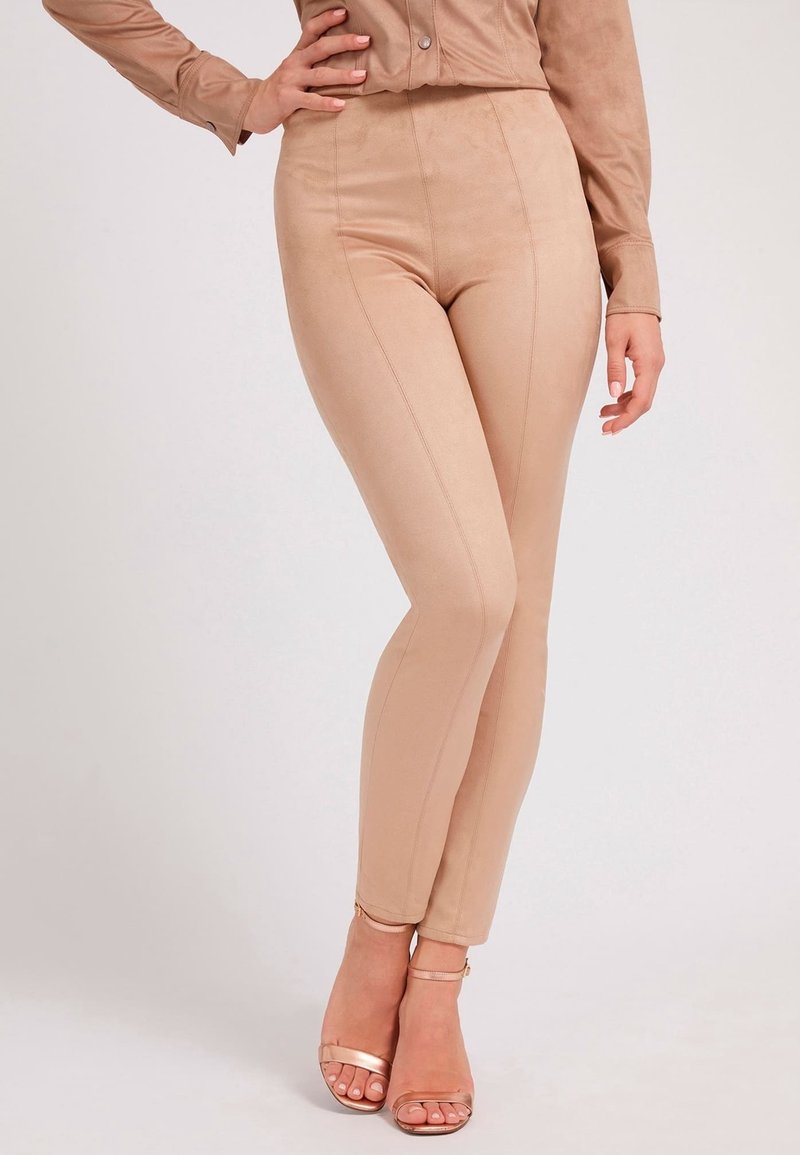 Leggings nude in suede sintetico con vita alta, texture liscia e dettagli di cucitura lungo la parte anteriore; abbinati a tacchi nude con cinturini.