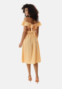 Yellow gingham dress with off-shoulder ruffles and a tied back, featuring a fitted waist and a midi-length skirt. Worn with orange heels.