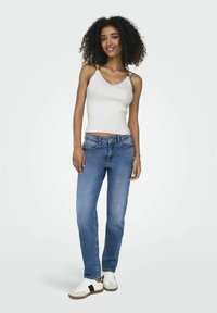 Witte geribde tanktop met verstelbare gouden schouderbandjes, gecombineerd met lichtblauwe rechte jeans en witte sneakers met zwarte accenten.