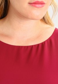 Haut bordeaux avec un col large et arrondi, et un drapé subtil autour du col. Tissu lisse et texturé, sans éléments métalliques visibles.