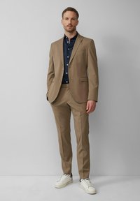 Beige pak met smalle revers, blazer met één knoop en vlakke broek. Gecombineerd met een donkerblauw shirt en witte sneakers. Glad textuur.