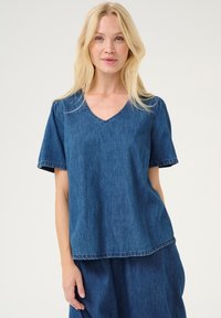 Zila denima krekls ar īsām piedurknēm un V veida kakla izgriezumu. Tas ir ar brīvu piegriezumu un neapstrādāts šuvums ap kakla izgriezumu un piedurknēm.