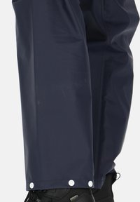 Marine waterdichte broek met een gladde textuur, drukknoopsluiting aan de onderkant en zwarte schoenen die gedeeltelijk zichtbaar zijn.