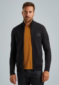 Giacca a zip nera in maglia con tessuto testurizzato, presenta un colletto alto e un badge con logo sul petto, indossata sopra una maglietta color senape.
