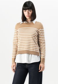 Braun gestreifter Pullover mit weißen Streifen, gerippte Bündchen und Saum, über einem weißen Hemd mit Nadelstreifen und Kragen getragen. Dunkle Jeans runden das Outfit ab.
