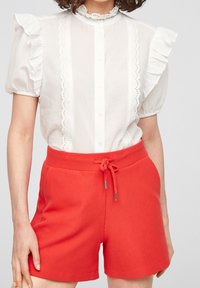 Witte blouse met ruches aan de mouwen en kantafwerking, gecombineerd met rode geribde shorts met een elastische tailleband en trekkoord.