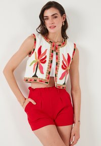 Débardeur blanc brodé avec des motifs floraux colorés, bordé d'un passepoil à motifs. Associé à un short plissé rouge.