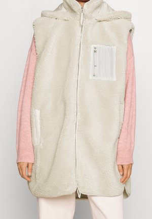 Veste sans manches en polaire couleur crème avec fermeture éclair frontale et poche poitrine zippée, portée sur un pull rose à manches longues.