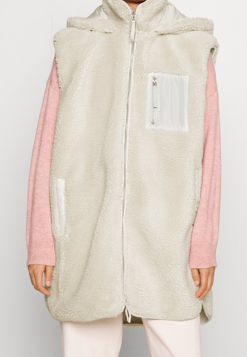 Veste sans manches en polaire couleur crème avec fermeture éclair frontale et poche poitrine zippée, portée sur un pull rose à manches longues.