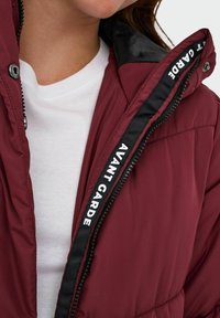 Burgundy puffer-jacka med svart, glänsande krage och kontrasterande vit text "AVANT GARDE" längs dragkedjan. Har en slät textur.