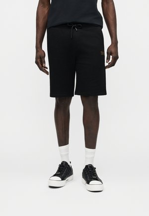 Férfi, fekete, térdig érő rövidnadrágban, fehér zoknival és fekete alacsony szárú tornacipőben, sima fehér háttér előtt állva.