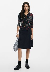 Must-õieline musta värvi nööpidega pluus pika varrukaga, kombineeritud põlvepikkuse tumeda seeliku ja mustade paksude kingadega. Käes hõbedane käekott.