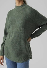 Personne portant un pull en tricot vert ample avec col et poignets côtelés, associé à un jean bleu, main posée sur la hanche.