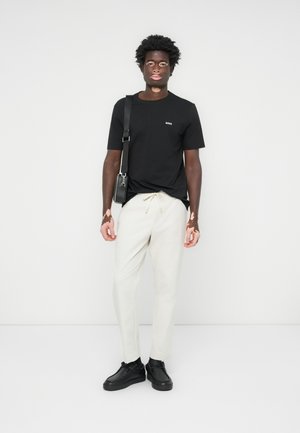 Schwarzes T-Shirt mit kleinem Logo, crèmefarbene Hose und schwarze Schuhe. Schwarze Umhängetasche und strukturiertes Haar. Stehend vor einem schlichten Hintergrund.