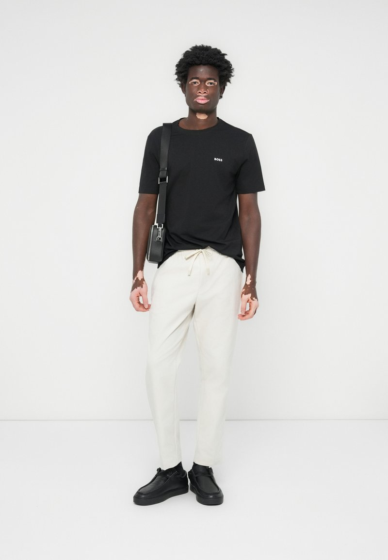 Zwarte t-shirt met een klein logo, crèmekleurige broek en zwarte schoenen. Zwarte schoudertas en gestructureerd haar. Staand tegen een effen achtergrond.