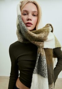 Uld tørklæde med et ternet mønster i nuancer af oliven, fløde og grå. Blød tekstur draperet om halsen, mod en neutral baggrund.