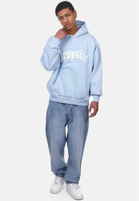 Hellblaue, übergroße Kapuzenjacke mit Fronttasche und weißem Branding, kombiniert mit hellen Jeans und weißen Sneakers. Lässiger, entspannter Schnitt.