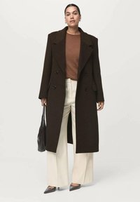 Abrigo largo de paño marrón, con doble botonadura, que presenta grandes solapas y botones. Lleva puesto un suéter marrón y unos pantalones de pierna ancha color crema.