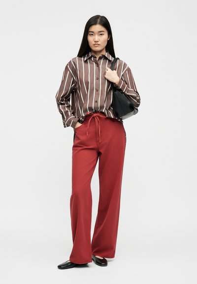 Νεαρή γυναίκα με καφέ και λευκό ριγέ πουκάμισο με κουμπιά, κόκκινο παντελόνι φαρδιάς γραμμής, μαύρες μπαλαρίνες, κρατώντας μαύρη τσάντα στον ώμο.