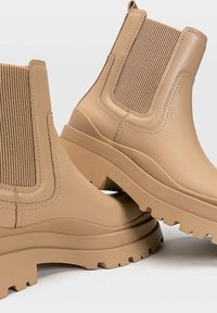 Bottines beige à talon mi-haut avec des côtés élastiques côtelés, une semelle épaisse et une finition lisse. Elles présentent un léger talon et des accents texturés pour une meilleure adhérence.