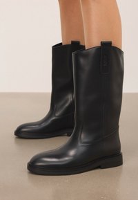 Stivali in pelle neri a metà polpaccio con punta arrotondata e linguette, indossati su gambe nude e in piedi su un pavimento beige liscio.