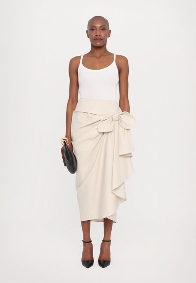 Jupe enveloppante beige avec une taille ajustée et un grand nœud, associée à un débardeur blanc. Les accessoires comprennent une pochette noire et des escarpins pointus.