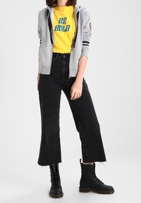 Grauer Kapuzenpullover mit Reißverschluss und schwarzen Streifen, gelbes T-Shirt mit "BE BOLD"-Aufdruck, schwarze gekürzte Jeans und schwarze Schnürstiefel.