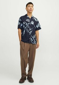 Camicia a maniche corte di colore blu navy con motivo floreale azzurro, chiusura con bottoni, abbinata a pantaloni larghi marroni e scarpe scure su uno sfondo neutro.