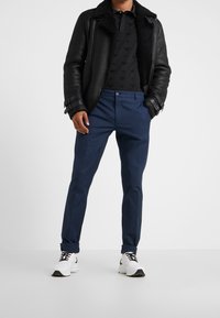 Černá kožená bunda s beránkovým límcem, černé vzorované polo tričko, modré kalhoty a bílé tenisky s černými akcenty.