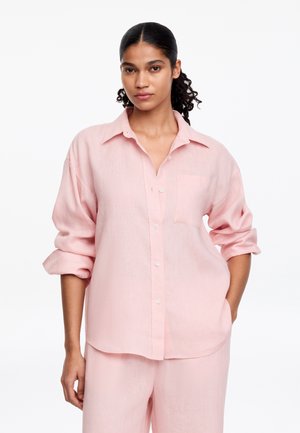 Femme aux cheveux bouclés attachés, portant une chemise ample rose clair avec des boutons et un pantalon assorti, debout avec une main dans la poche.