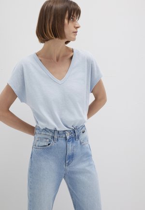 Helles blaues V-Ausschnitt-T-Shirt mit kurzen Ärmeln, kombiniert mit hochgeschnittenen hellen Jeans. Der Hintergrund ist neutral und schlicht.