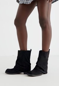 Botas slouch pretas feitas de camurça, com bico arredondado e detalhes texturizados sutis, equipadas com uma sola de borracha flexível.