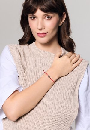 Czerwona bransoletka sznurkowa z ozdobnym srebnym charms'em, noszona na nadgarstku modelki w zestawieniu z jasnym swetrem. Elegancki, schludny design.