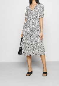 Biała sukienka z czarnym abstrakcyjnym wzorem, krótkie rękawy, dekolt w serek, szew w talii i falbaniasta spódnica. Połączona z czarnymi sandałami na słupku i torebką.