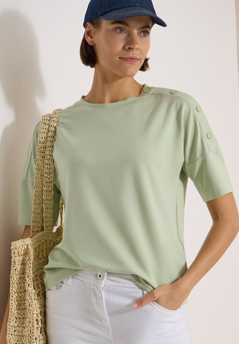 Femme portant une chemise vert clair avec des boutons sur l'épaule, pantalon blanc, casquette bleu foncé, tenant un sac beige tissé sur l'épaule.