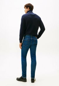 Uomo in dolcevita blu scuro, jeans slim-fit blu e scarpe eleganti nere, in piedi con una mano sulla vita e rivolto di spalle.