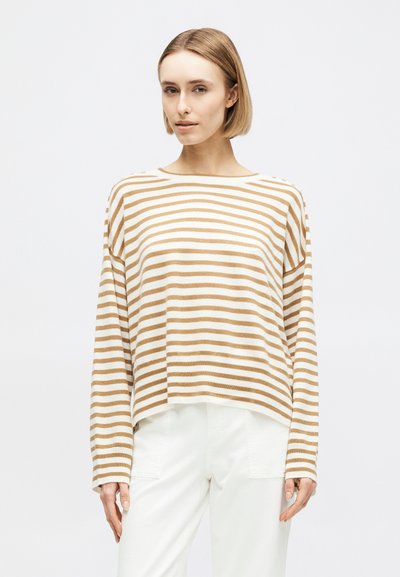 Femme portant un pull à manches longues beige et blanc à rayures horizontales et un pantalon blanc, debout devant un fond blanc uni.