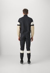 Tuta nera con accenti crema e navy, caratterizzata da un colletto alto, polsini elastici e gambe aderenti con ampie strisce crema sulla parte inferiore delle gambe.