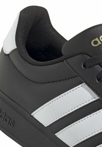 Gros plan sur une chaussure de sport Adidas noire et blanche montrant un col rembourré, trois bandes blanches et le logo Adidas doré sur la languette.