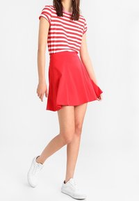 Roter, ausgestellter Rock kombiniert mit einem rot-weiß gestreiften Kurzarmoberteil, getragen mit weißen Sneakers. Glatte Textiloberfläche, lässiges Design.