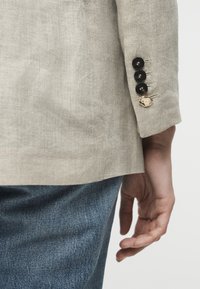 Μανίκι μπεζ λινό σακάκι με τέσσερα κουμπιά, ένα χρυσό, φορεμένο με μπλε τζιν τζιν, το χέρι χαλαρό στο πλάι.