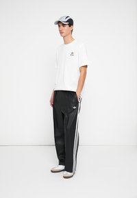 White cotton t-shirt with small black logo, black track pants with white stripes, grey and black cap, and cream sneakers with brown accents.