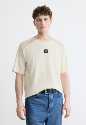 Młody mężczyzna z kręconymi włosami, ubrany w beżową koszulkę z krótkim rękawem z czarną naszywką z logo oraz niebieskie dżinsy, na białym tle.